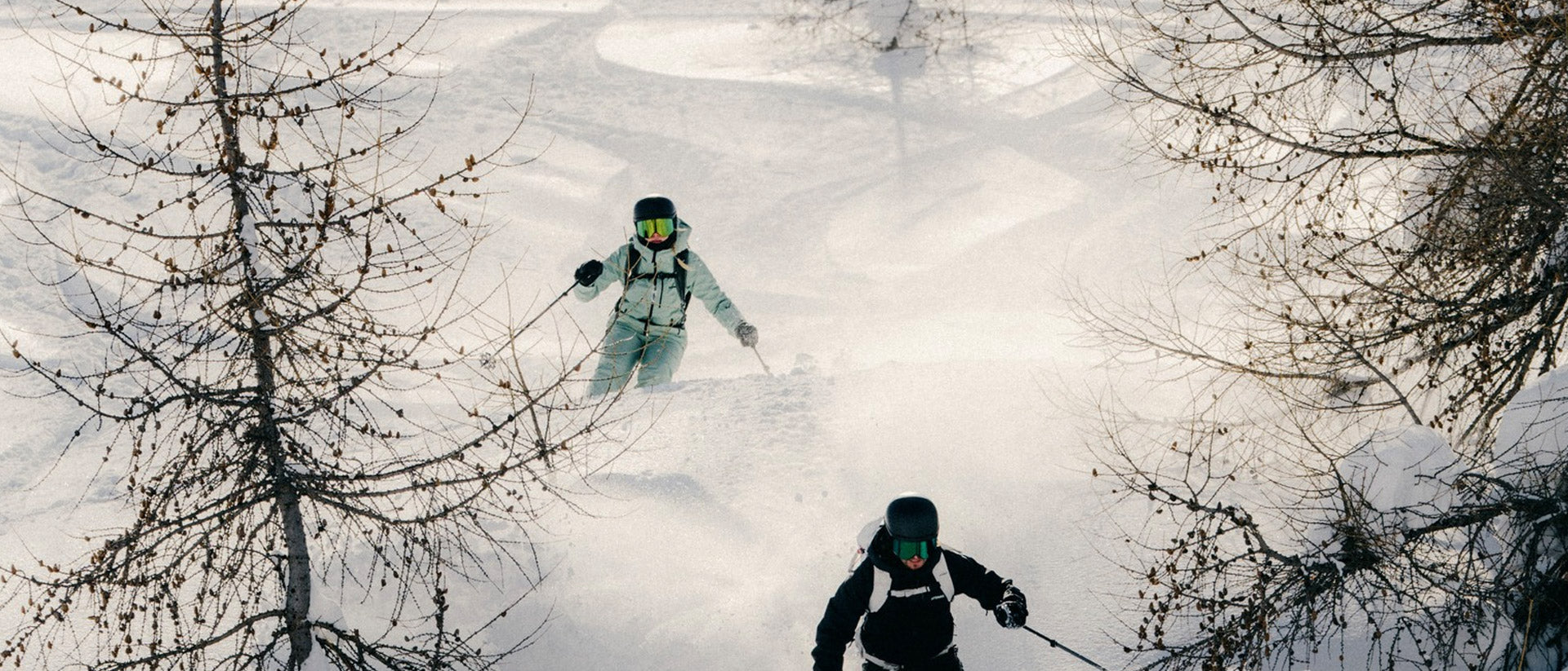 Two skiers navigating a snowy landscape with trees wearing Atomic Revent visor ski helmets
