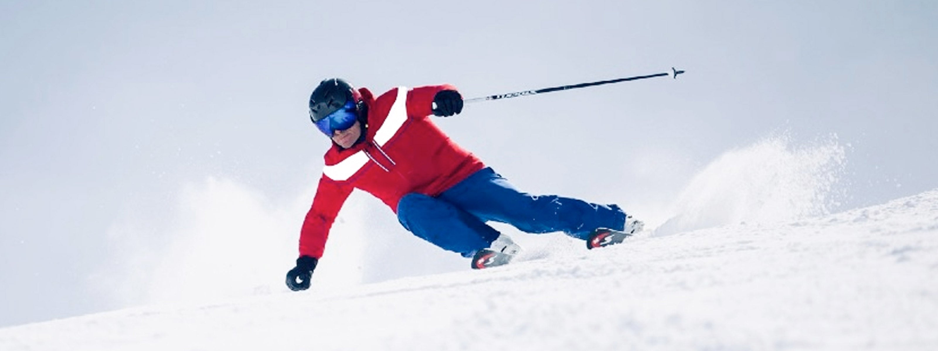 Skier in red jacket, blue pants and black carbon ski helmet skiing down a snowy slope with a clear sky.