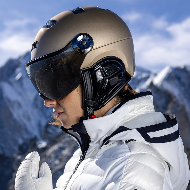 Person wearing a gold visor ski helmet from Kask and white jacket with mountains in the background